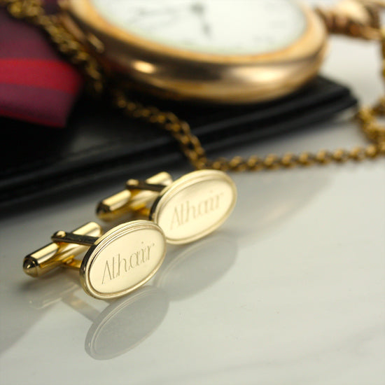 Gold cufflinks with 'Athair' engraving on a reflective surface, with a pocket watch in the background.